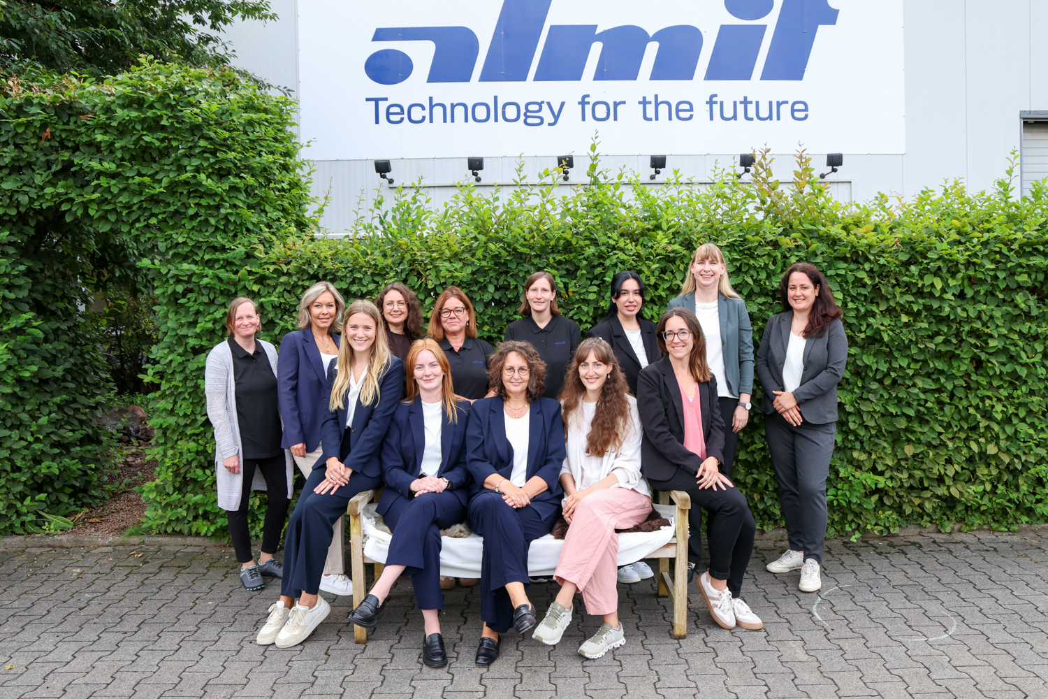 Das Verwaltungsteam der Almit GmbH vor einer Hecke, dahinter die Fassade mit dem Schriftzug „almit – Technology for the future“