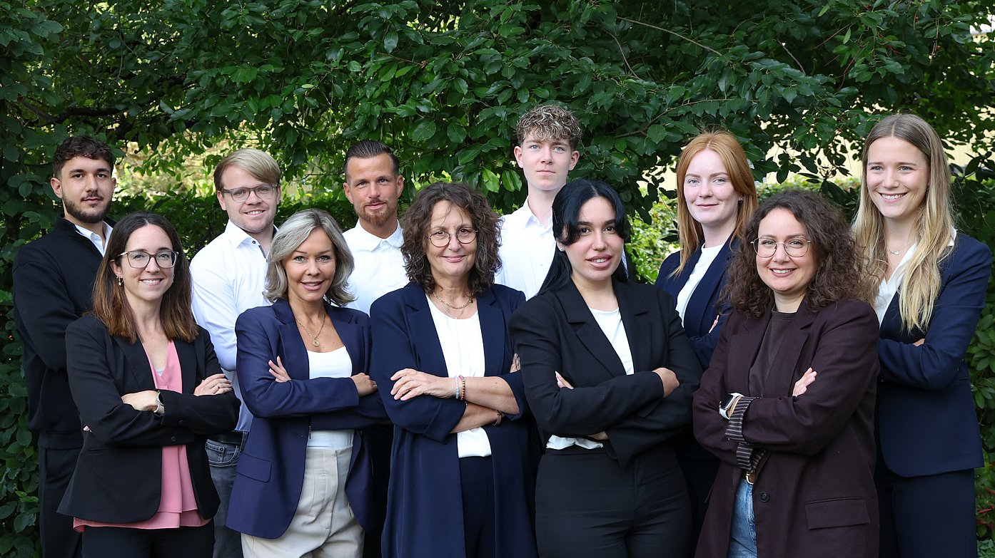 Das Team Verwaltung von Almit, elf Personen in Business-Kleidung vor einer Hecke